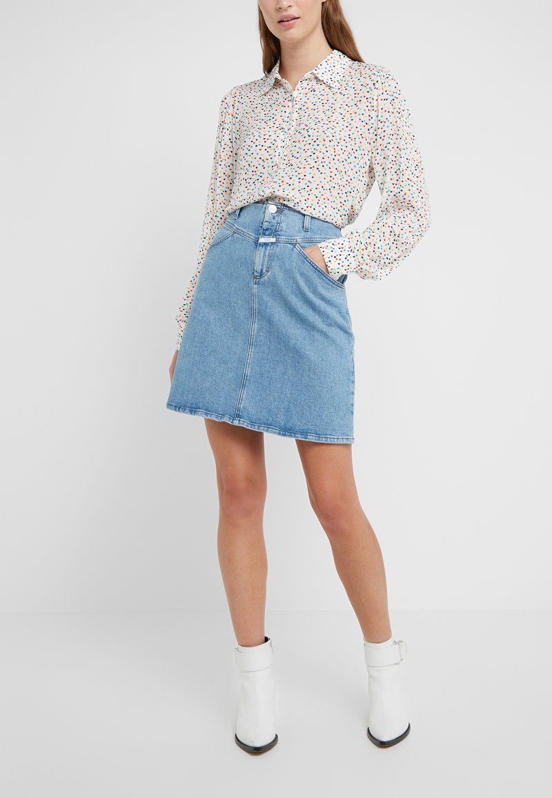 Femme portant un chemisier blanc à pois multicolores, une jupe en jean bleue et des bottines blanches, debout avec une main dans la poche.
