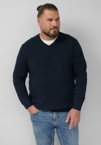 Granatowy sweter z dekoltem w serek, wykonany z miękkiego materiału, z długimi rękawami i ściągaczami, w połączeniu z jasnoniebieskimi dżinsami.