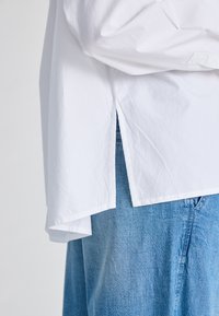 Camicia bianca in cotone con orlo spaccato, sovrapposta a un jeans in denim blu chiaro. Presenta una vestibilità rilassata e un design minimalista.