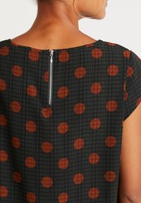 Vêtement noir avec de grands pois marron et un motif en grille texturé. Doté d'une courte manche à capuchon et d'une fermeture éclair visible au dos.