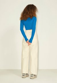 Langærmet blå crop top, cremefarvede bukser med vide ben og grå sneakers. Synlige syninger på bukserne; let tekstureret stof. Hænderne bag ryggen.