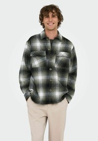 Camisa de franela en patrón de cuadros en negro, gris y blanco, con dos bolsillos en el pecho y botonadura frontal, llevada sobre una camiseta oscura, combinada con pantalones beige.