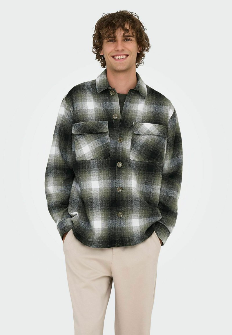 Camisa de franela en patrón de cuadros en negro, gris y blanco, con dos bolsillos en el pecho y botonadura frontal, llevada sobre una camiseta oscura, combinada con pantalones beige.