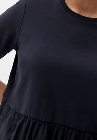 T-shirt bleu marine foncé à manches courtes, confectionné en tissu lisse. Coupe légèrement évasée avec une taille froncée, créant une silhouette décontractée.