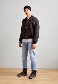 Schwarze Bomberjacke mit silbernen Druckknöpfen, grauer Ripp-Strickpullover, hellblaue Jeans und schwarze High-Top-Sneaker mit grauen Sohlen.