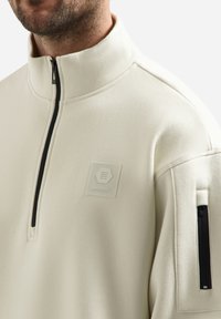 Crèmekleurige fleece trui met een hoge kraag, kwart-rits ontwerp en een ritszak op de linkermouw. Voorzien van een gestructureerde logo patch.