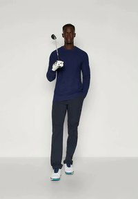 Navy long-sleeve shirt paired with dark pants and white golf shoes. A golf club is held in the left hand, with a white glove on the right hand.