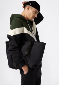 Uomo che indossa una felpa con cappuccio nera, bianca e verde scuro e un berretto nero, mentre porta una grande borsa a tracolla nera sulla spalla, con una mano in tasca.