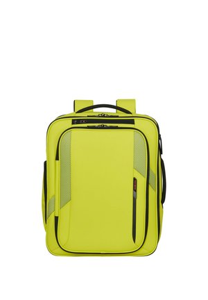 Hellgelber quadratischer Rucksack mit schwarzen Reißverschlüssen, Fronttaschen, Netzdetails, Tragegriff oben und gepolsterten Schultergurten.