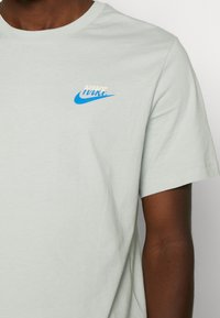 Light gray cotton t-shirt with short sleeves. Features a small blue Nike logo on the left chest and a rounded neckline.
