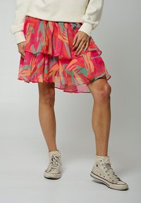 Jupe empilée jusqu'aux genoux en rose avec des motifs abstraits en vert et orange. Tissu léger et transparent avec des volants. Portée avec des baskets blanches.