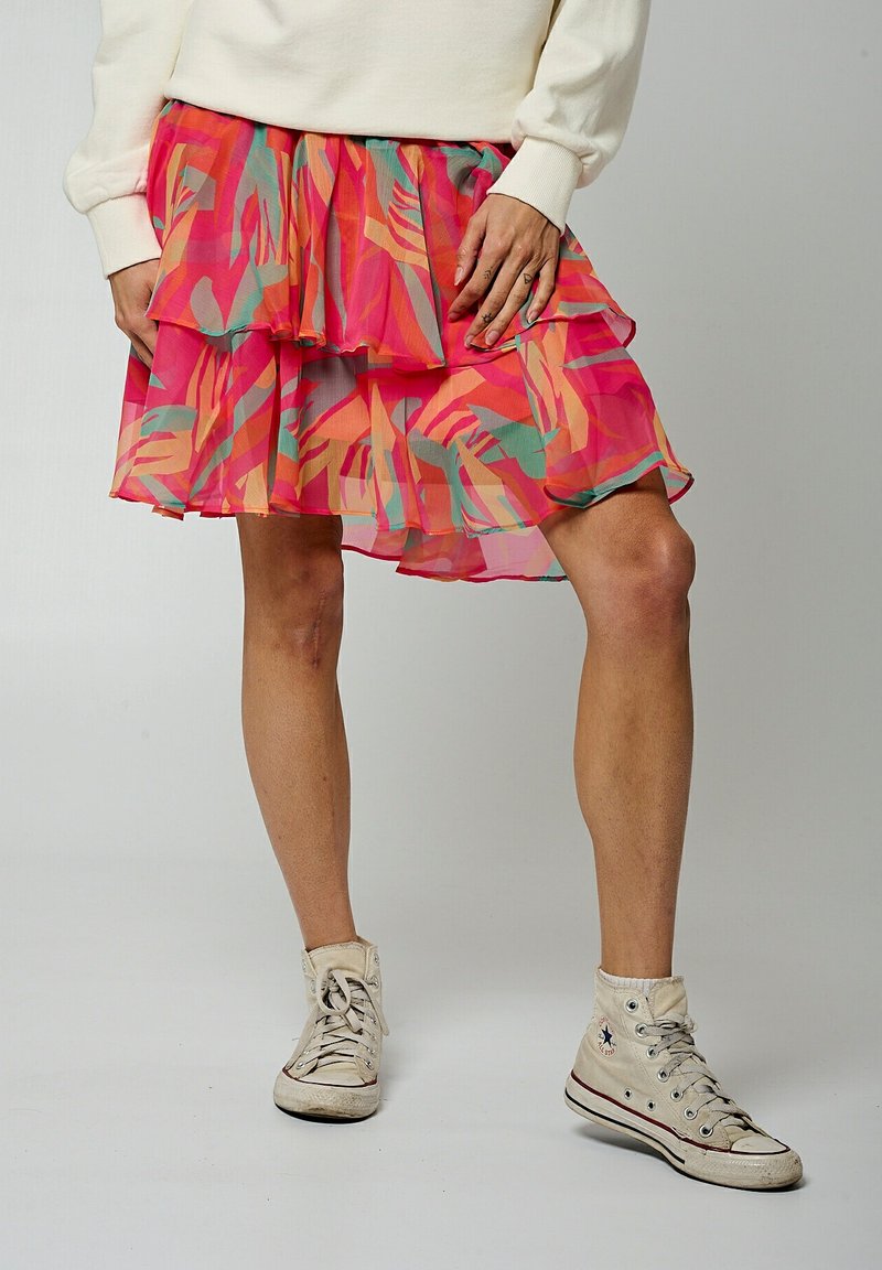 Jupe empilée jusqu'aux genoux en rose avec des motifs abstraits en vert et orange. Tissu léger et transparent avec des volants. Portée avec des baskets blanches.