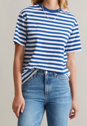 Vrouw draagt een blauw-wit gestreept T-shirt met korte mouwen, ingestopt in high waist blauwe jeans met een zilveren ketting.