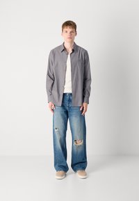Camisa de botones a rayas gris y blanca sobre una camiseta blanca, combinada con jeans de pierna ancha de color azul claro que presentan parches desgastados. Zapatillas de deporte beige.