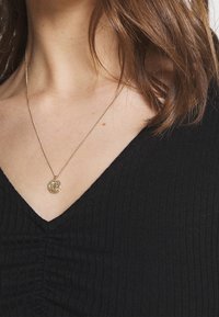 Kvinna med axellångt brunt hår som bär ett guldhänge och en svart V-ringad ribbad topp.