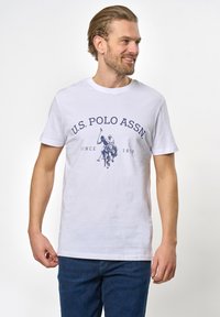 Balta medvilninė marškinėliai su tamsiai mėlynu U.S. Polo Assn. logotipu ir dviejų polo žaidėjų grafika. Trumpomis rankovėmis ir apvalia kaklo iškirpte.