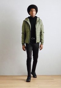 Veste matelassée verte avec fermeture éclair, portée sur un col roulé noir. Le modèle porte un pantalon noir ajusté et des baskets noires. Sol en bois lisse.