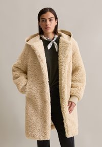 Crème kleurige, fuzzy jas met een ontspannen pasvorm, een oversized capuchon en geribbelde manchetten. Gedragen over een donkere top en aangevuld met een gestreepte sjaal.