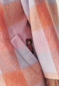 Villane mantel roosa ja oranži gradatsiooniga ruudulises mustris; varustatud taskuga, millel on kanga peal karu illustratsioon. Pehme, tekstuuriga viimistlus.