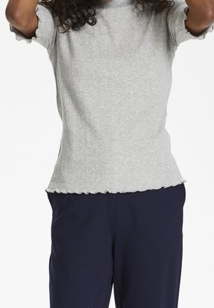 Osoba w jasnoszarej, żeberkowanej bluzce z krótkim rękawem i falbanką u dołu oraz granatowych spodniach, z uniesionymi ramionami nad głową.