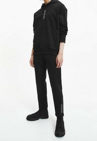 Felpa nera con cappuccio, tasca frontale e dettaglio testo verticale; abbinata a pantaloni neri e stivali neri stringati, tutti con design aderente.