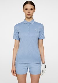 Vaaleansininen polo paita, jossa on tekstuuri, lyhyet hihat ja kolme nappia. Yhdistetty vaaleansinisiin shortseihin. Valkoinen logo rinnassa.