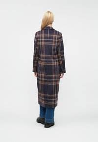 Manteau en mélange de laine arborant un motif à carreaux navy et marron, à manches longues, avec un col à revers et une coupe mi-longue, associé à un jean foncé et des bottes.