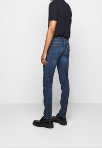 Jeans in denim skinny-fit di colore blu scuro, con cinque tasche e dettagli di cucitura sottili, abbinati a scarpe in pelle nera.