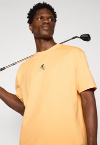Oransje bomull t-skjorte med korte ermer og rund halslinning. Har en liten svart logo på brystet. Golfklynge hviler på skulderen.