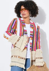Cardigan colorato con motivo a zigzag e floreale, maniche in pizzo color crema e dettagli a frange. Indossato sopra una camicia bianca.