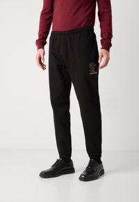 Zwarte joggingsbroek met een elastische tailleband, voorzien van een subtiele Lacoste-logo in goud op de linker dij. Draagt zwarte schoenen.