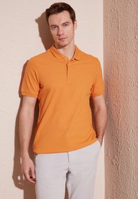 Oranžové polo tričko s klasickým límečkem, krátkými rukávy a zapínáním na tři knoflíky; v kombinaci se světlými šedými kalhotami. Hladká textura látky.