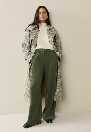 Long plaid coat with a light brown and blue pattern, cream turtleneck, and wide green corduroy pants. Black footwear completes the outfit.