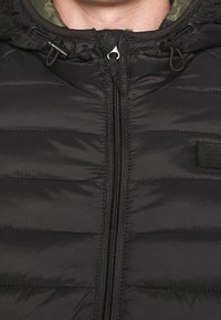 Doudoune noire avec des segments matelassés horizontaux, fermeture éclair à l'avant et capuche à cordon de serrage. Texture lisse avec fini mat. Étiquette de marque visible.