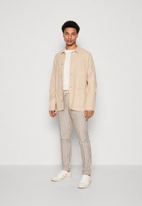 Giacca in lino beige, t-shirt bianca a girocollo, pantaloni a righe beige e marroni, e sneakers bianche con accenti beige.