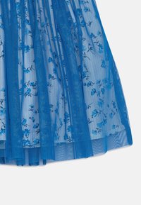 Modrá květinová látka s průhledným modrým síťovaným překryvem, ukazující záhyby a texturu, umístěná na bílém pozadí.