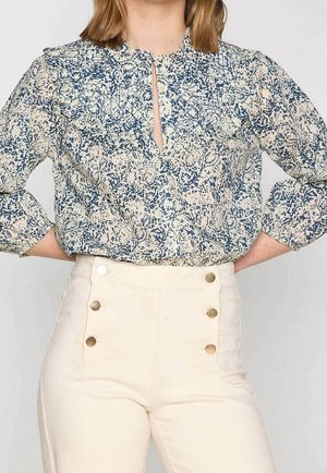 Blomsterbluse i creme og marineblå med flæsekrave og en forreste nøglehul. Parret med højtaljede cremebukser med gyldne knapdekorationer.