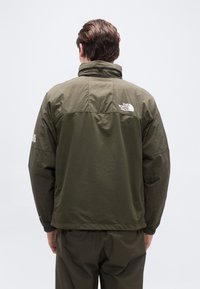 Άνδρας με μπουφάν The North Face σε ελιά πράσινη απόχρωση, με το λογότυπο στην άνω πλάτη και το μανίκι, απεικονίζεται από πίσω σε απλό φόντο.