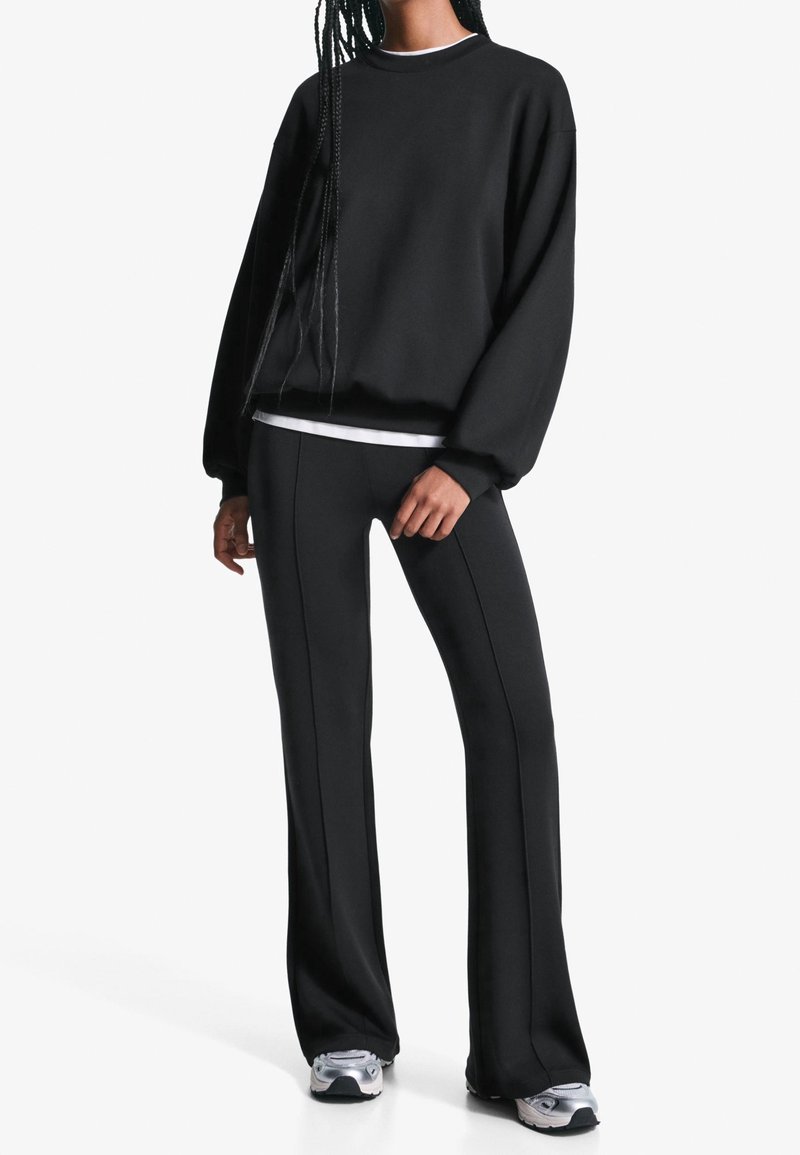 Femme portant un sweat-shirt noir à manches longues, un pantalon noir à jambes larges assorti, un sous-vêtement blanc et des baskets argentées, debout sur un fond blanc.