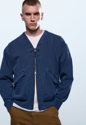 Νεαρός άνδρας με κοντά μαλλιά που φοράει μπλε μπουφάν με φερμουάρ πάνω από λευκό πουκάμισο και καφέ παντελόνι, στέκεται με τα χέρια στις τσέπες.