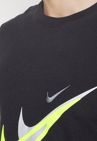 Černé tričko Nike s kulatým výstřihem, na přední straně s kovově stříbrnými a neonově žlutými logy Swoosh. Hladká bavlněná textilie.