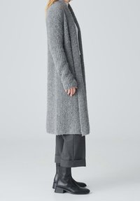 Persona vistiendo un largo cárdigan gris texturizado, pantalones oscuros cortos y botas negras hasta el tobillo, frente a un fondo gris claro.