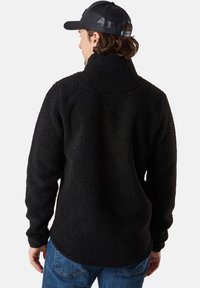 Svart fleece-tröja med hög krage, strukturerad yta, långa ärmar och en lätt rundad nederkant, bärs över blå jeans.