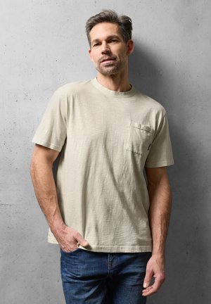 Homme aux cheveux gris courts portant un t-shirt beige à manches courtes avec une poche et un jean bleu, debout devant un mur en béton gris.