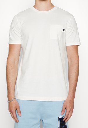 Mężczyzna w prostym białym t-shircie z krótkim rękawem i małą kieszonką na piersi, w połączeniu z jasnoniebieskimi szortami ozdobionymi dużą naszywką w kształcie litery K.