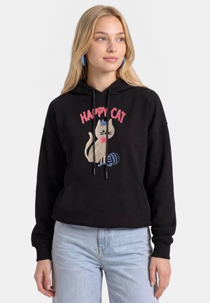 Giovane donna che indossa una felpa nera con cappuccio con un gatto dei cartoni animati, un gomitolo di lana e il testo "Happy Cat,", abbinata a jeans azzurri e orecchini a cerchio blu.