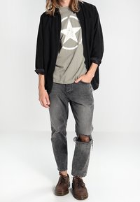 Conjunto casual que incluye una camiseta negra, una camiseta gráfica verde oliva con una estrella, jeans grises desgastados y zapatos marrones.