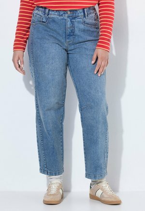 Persoon draagt blauwe mid-rise jeans, beige sneakers met witte en groene strepen, witte sokken en een rood shirt met lange mouwen met oranje strepen.