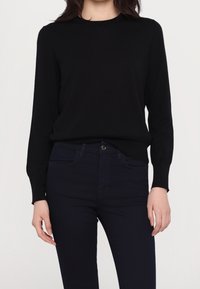 Kvinna klädd i en figurnära svart långärmad tröja och mörkblå högmidjade skinny jeans mot en enkel ljus bakgrund.