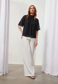 Blusa negra de manga corta con escote plisado, combinada con pantalones blancos de pierna ancha. Textura suave con acentos translúcidos y diseño minimalista.